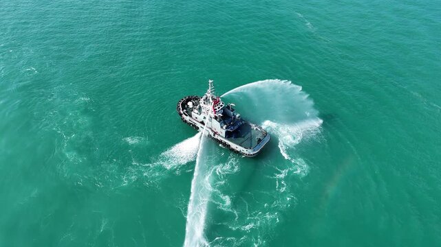  tugboat operating a powerful water cannon, spraying water in a busy shipyard harbor with large industrial cranes and cargo ships in the background. Maritime operations