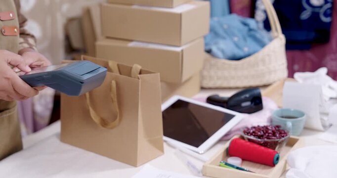Retired asian woman small business holds POS terminal during checkout, craft seller at home reviews inventory management desk, sorts handmade shibori fabric order, prepares e-commerce parcel packing.