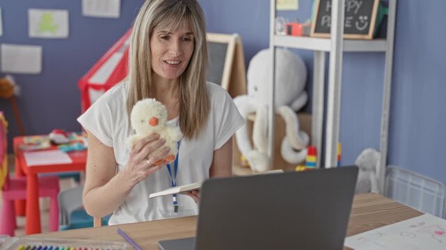 Woman teacher holds stuffed duck and smiles while reading from a book to a laptop in a classroom building; warmth remote learning.