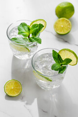 Fototapeta premium A fresh mojito with lime, mint and ice in glasses. Citrus lemonade or cocktail on a marble table with shadows.