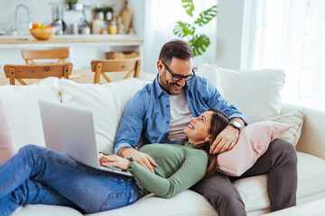 Obraz premium Smiling Couple Relaxing on Sofa at Home Using Laptop Together in Bright Living Room