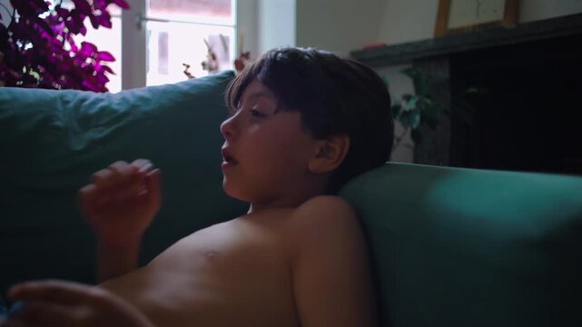 Young boy lying on couch picking nose and wiping face showing candid everyday behavior during relaxed moment at home