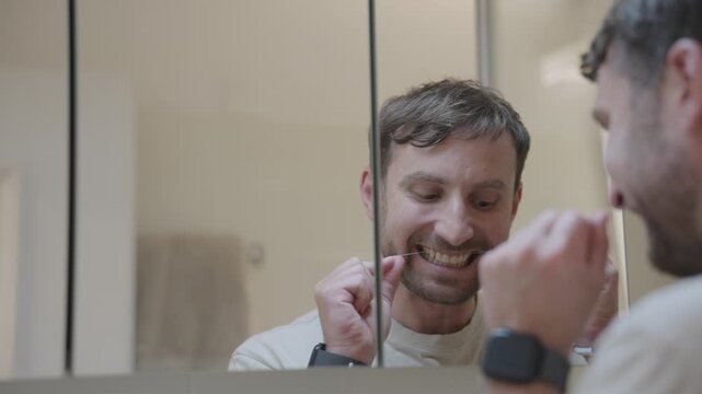Man looking at his reflection while taking care of his oral hygiene with dental floss at home