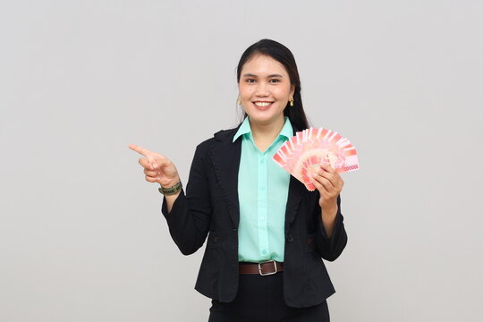 A happy and surprised young businesswoman receives rupiah cash, pointing to the side in an empty space, standing isolated on a white background.