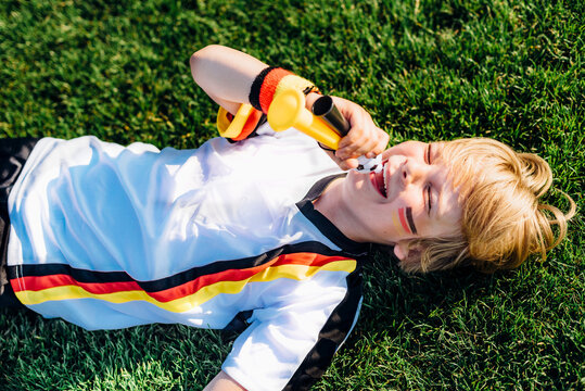 Boy in German soccer shirt lying on grass, blowing horns