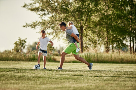 Man giving piggyback ride to daughter by son playing soccer at sports field