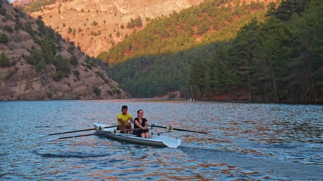 Cinematic shot of two rowers moving across a blue lake beside rocky cliffs. Dynamic outdoor sports scene with freshwater, rowing motion, teamwork and natural landscape.