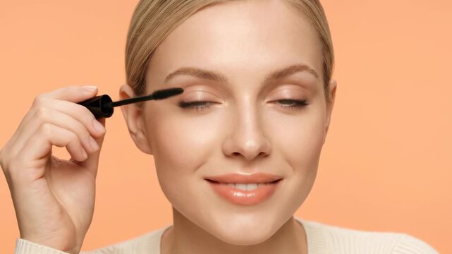 A smiling, attractive young Caucasian woman with blonde hair and fair skin gently applies black mascara to her eyelashes with her eyes closed. The scene is set against a clean, solid peach background,