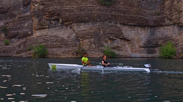 Cinematic shot of two rowers moving across a blue lake beside rocky cliffs. Dynamic outdoor sports scene with freshwater, rowing motion, teamwork and natural landscape.