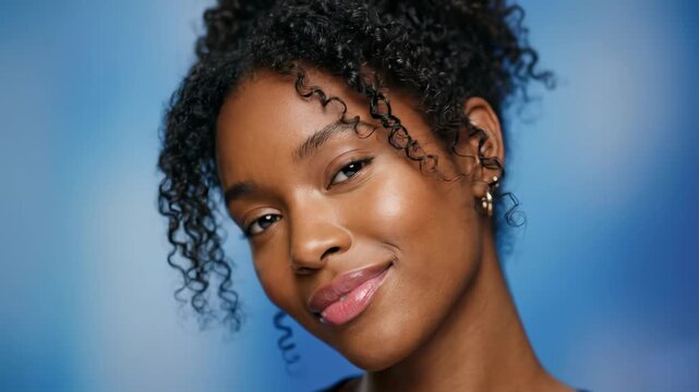 A dark-skinned young woman with dark curly hair in a top poses flirtatiously and smiles charmingly at the camera against a blue background. Suitable for beauty product ads, cosmetics, or lifestyle bra
