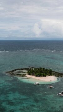 Small island with boats anchored nearby, turquoise sea and cloudy sky stretching across the horizon. Guyam Island. Siargao, Philippines.