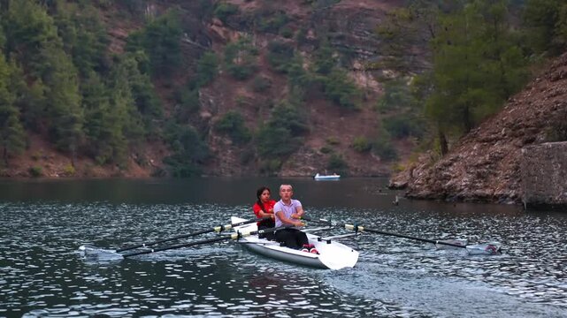 Cinematic close view of two rowers moving across a lake at sunset near a rocky shore. Peaceful outdoor sports scene with warm reflections, freshwater and natural landscape.