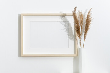 Horizontal blank photo frame mockup on white wall next to modern white vase holding dried pampas grass, styled in a bright minimalist interior © nikavera