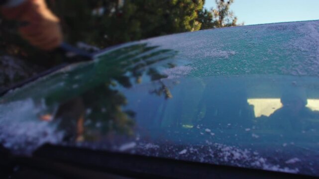 Hand scraping ice from car windshield with glove on cold winter morning, vehicle parked in snowy mountain landscape under bright sunlight