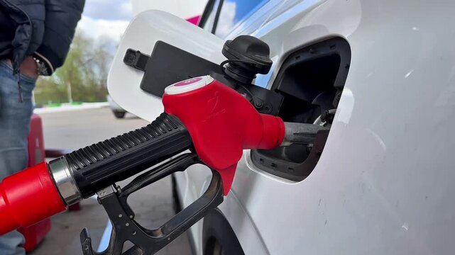 Video. A red pistol is inserted into the gas tank of a white car, with a man in the background.