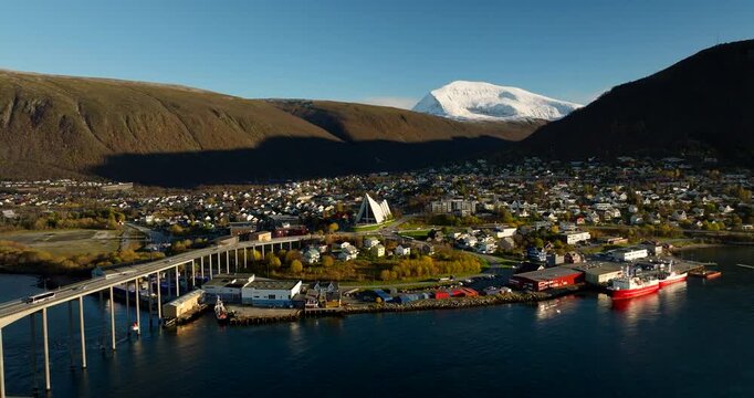 Tromso city, bridge, and Arctic Cathedral with snow capped mountains during sunny late autumn day, Norway. Aerial backward