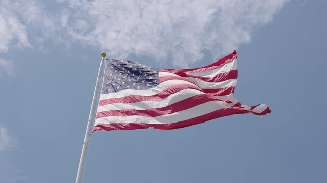 A large, front lit American flag blowing in the wind in slow motion with a partly cloudy blue sky in the background. Low angle.