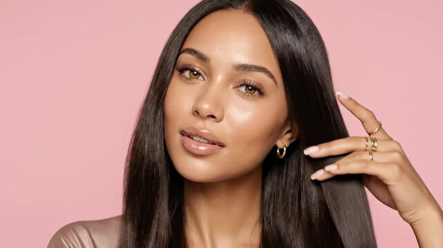 A gorgeous young Black woman with warm brown skin and long, glossy dark brown hair smiles beautifully while gently stroking and fixing her locks. The video is shot against a soft, solid pink studio ba