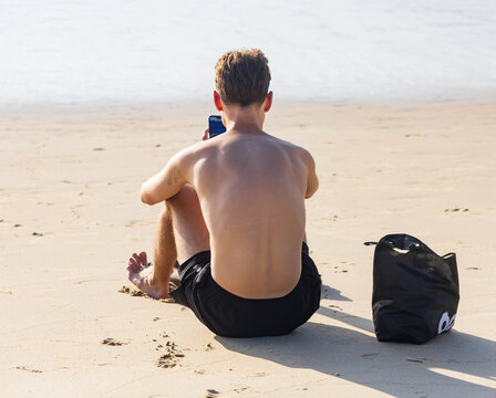 Man sits bareback on sandy beach, holding phone