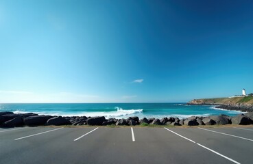 Fototapeta premium Empty asphalt parking area overlooks ocean waves crashing on rocky shore. Lighthouse stands on distant cliff under clear blue sky. Coastal landscape invites road trip travel.