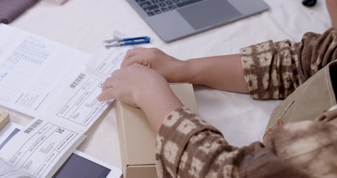 Senior asian entrepreneur peels shipping label, aligns barcode sticker on carton lid, checks invoice and receipt list, prepares parcel packaging station for delivery workflow in home studio.