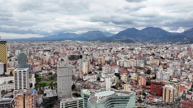 Aerial perspective flying forward and rising above Tirana, Albania, revealing modern skyscrapers like TID Tower, urban architecture, and a majestic mountain backdrop