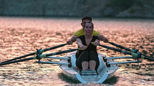 Close cinematic view of two rowers gliding across a calm lake surrounded by rocky hills and pine trees during sunset. Peaceful outdoor sports scene with teamwork and motion on freshwater.