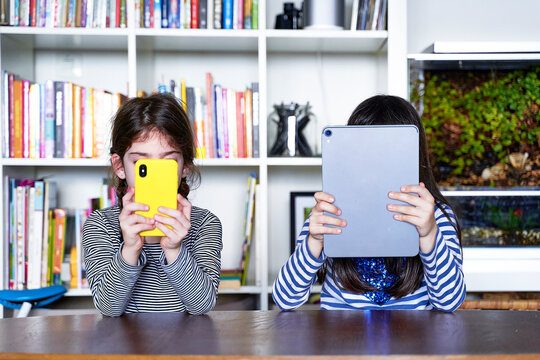 Two sisters sitting side by side at table in the living room using electronic devices
