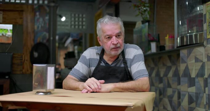 Older caf&eacute; owner sits at table with distant gaze conveying concern uncertainty and emotional strain as small business faces ongoing financial hardship