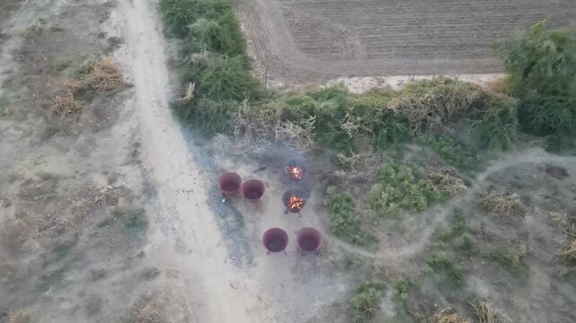 Aerial drone footage of traditional biochar and charcoal production using metal kilns in a rural countryside. Features biomass carbonization, firewood burning, smoke, sustainable energy, and rural ind
