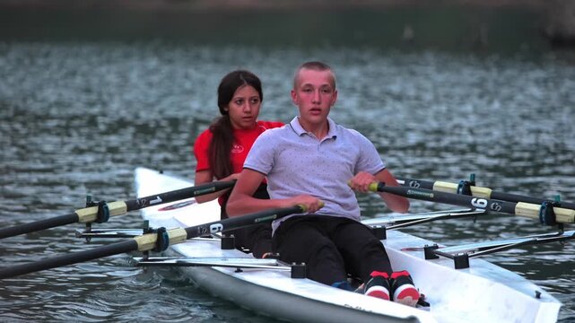 Close cinematic view of two rowers gliding across a calm lake surrounded by rocky hills and pine trees during sunset. Peaceful outdoor sports scene with teamwork and motion on freshwater.