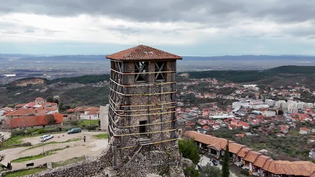Aerial camera circles counterclockwise around the historic stone clock tower under restoration, revealing the castle walls, Skanderbeg Museum, and sweeping valley panorama of Kruj&euml;, Albania