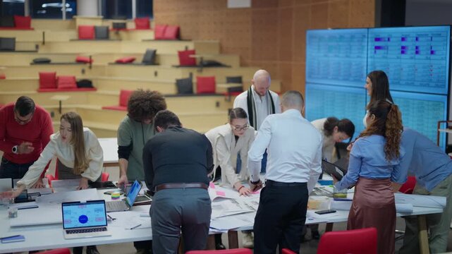 A group of colleagues actively brainstorm and review documents around a large table. Diverse employees engage in collaborative teamwork within a contemporary office setting.