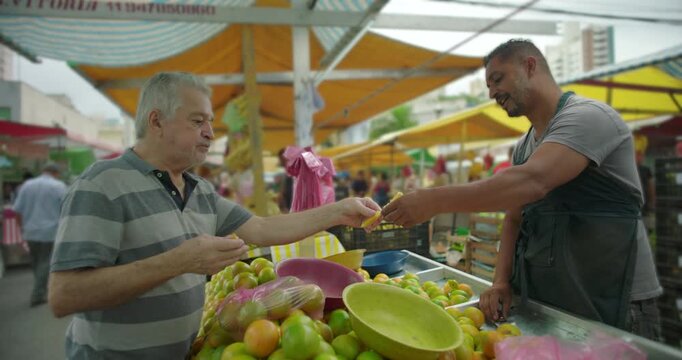 Hispanic street vendor handing fruit sample to elderly customer at produce stall showing care transparency and familiar relationship in local street food trade