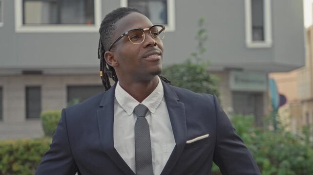 Man in tailored suit adjusts glasses while smiling on a street, tie and pocket square visible, dreadlocks and beard present; confidence ambition success.