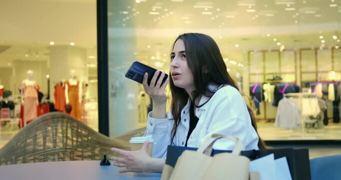 Woman recording voice message at mall cafe table, holding phone close to mouth, shopping bags beside, reflective storefront backdrop, thoughtful tone, casual outfit, coffee to go on table