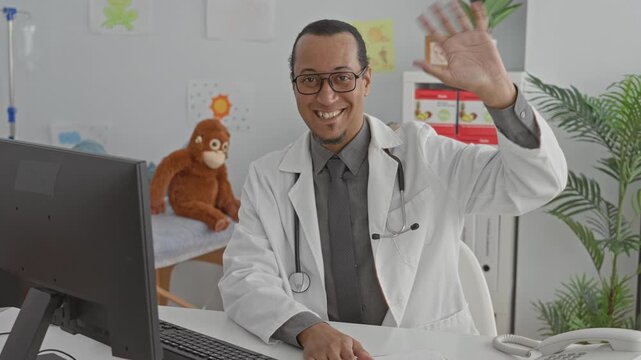Man doctor with stethoscope waving hand at computer in pediatric exam building with stuffed toy and plant visible; friendly telehealth greeting.