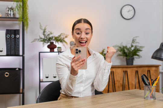 Young businesswoman at home office types on smartphone sees app game victory and smiles with joy. Freelancer girl at table raises fist in victory sends quick reply to message and celebrates good news.