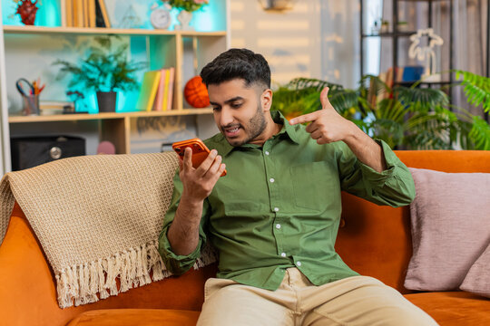 Indian man on home sofa speaks on smartphone speaker call with stop gesture and upset look in tense argument. Arabian guy on home sofa reacts with stress and anger in loud phone dispute indoors alone