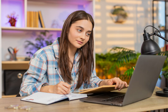 Young woman on home table studying with laptop and book while writing notes for online education at home. Adult girl on home table reads book and writes ideas with focused mood in living room indoors