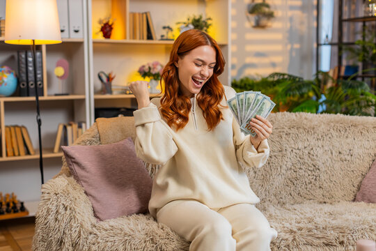 Young woman on home sofa counts cash with smile and checks amount while dreaming of purchase and travel. Adult girl on home sofa holds banknotes and enjoys thoughts of savings income and real success.