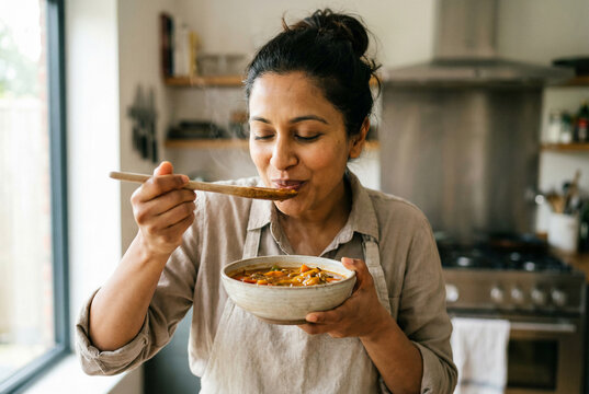 Indian woman savoring aroma of homemade vegetable soup in bright modern kitchen wearing apron with wooden spoon