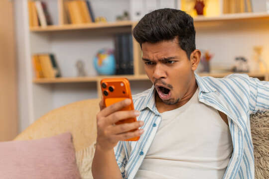 Indian man on home sofa reads bad news on smartphone and feels shock and disappointment during quiet home break. Arabian guy on home sofa reacts to negative message with raised brows and sad stress.