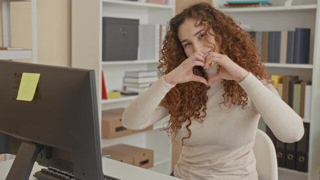 Young woman at computer desk forms heart with hands while smiling in building office; affection connection.