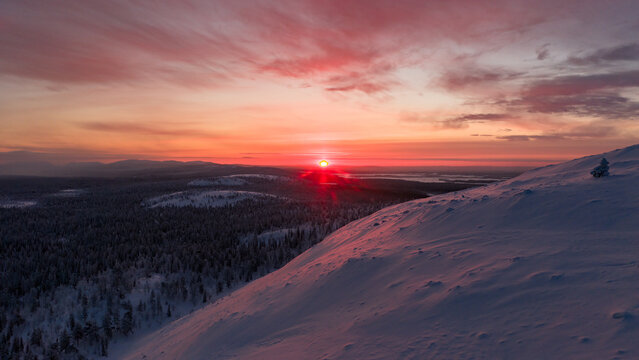 Aerial view of Luosto fell with snow-covered slopes and vast taiga forest under a vibrant red sunset sky Sodankyla, Lapland, Finland.