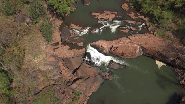 Drone shot descends over Tad Champee Waterfall north of Pakse, tilting up to reveal upstream rock pools, forest and flowing water in Laos.