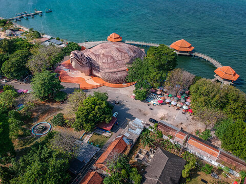 Aerial view of the giant turtle-shaped building of Kura-Kura Ocean Park on the coastline with wooden walkways and gazebos over turquoise water in Jepara, Central Java, Indonesia.