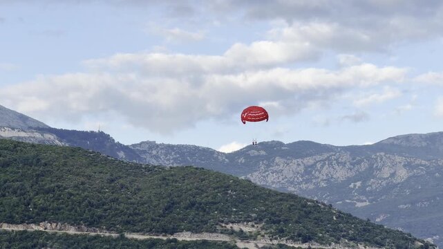 A red parasail with two people descends slowly against a backdrop of green mountains and a cloudy blue sky in Budva, Montenegro