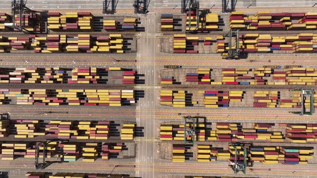 Containers are arranged in stacks at a busy port. Equipment is moving containers. Trucks are loading goods for transportation. Operations highlight the complexity of international trade.