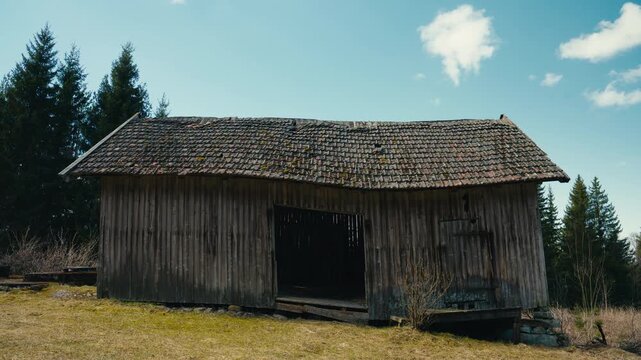 An old wooden barn stands alone at the edge of a forest, with worn timber, a sagging roof, and open doorway giving the rural scene a quiet and timeworn character.